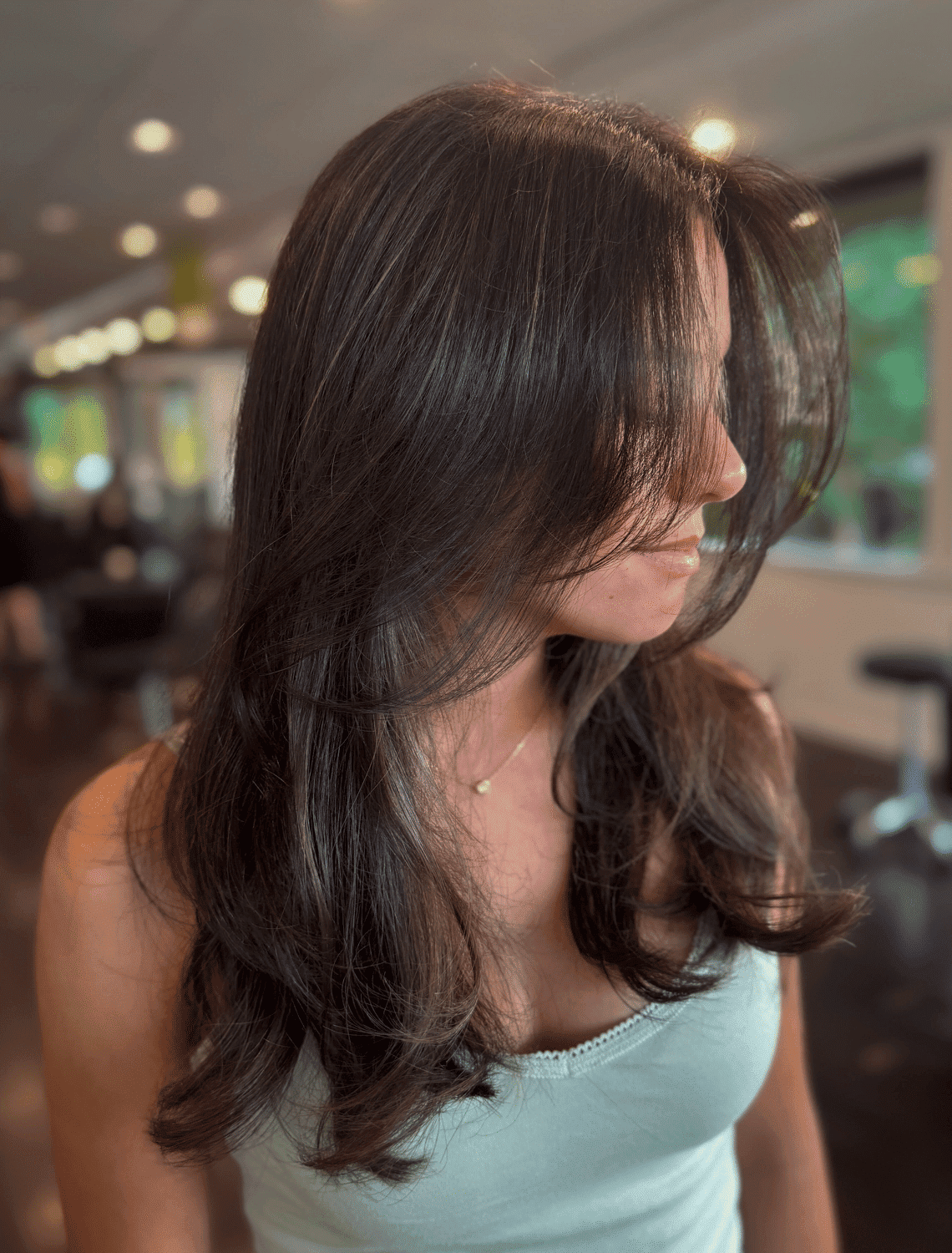 Woman with long, layered dark hair in a salon, wearing a light blue tank top.