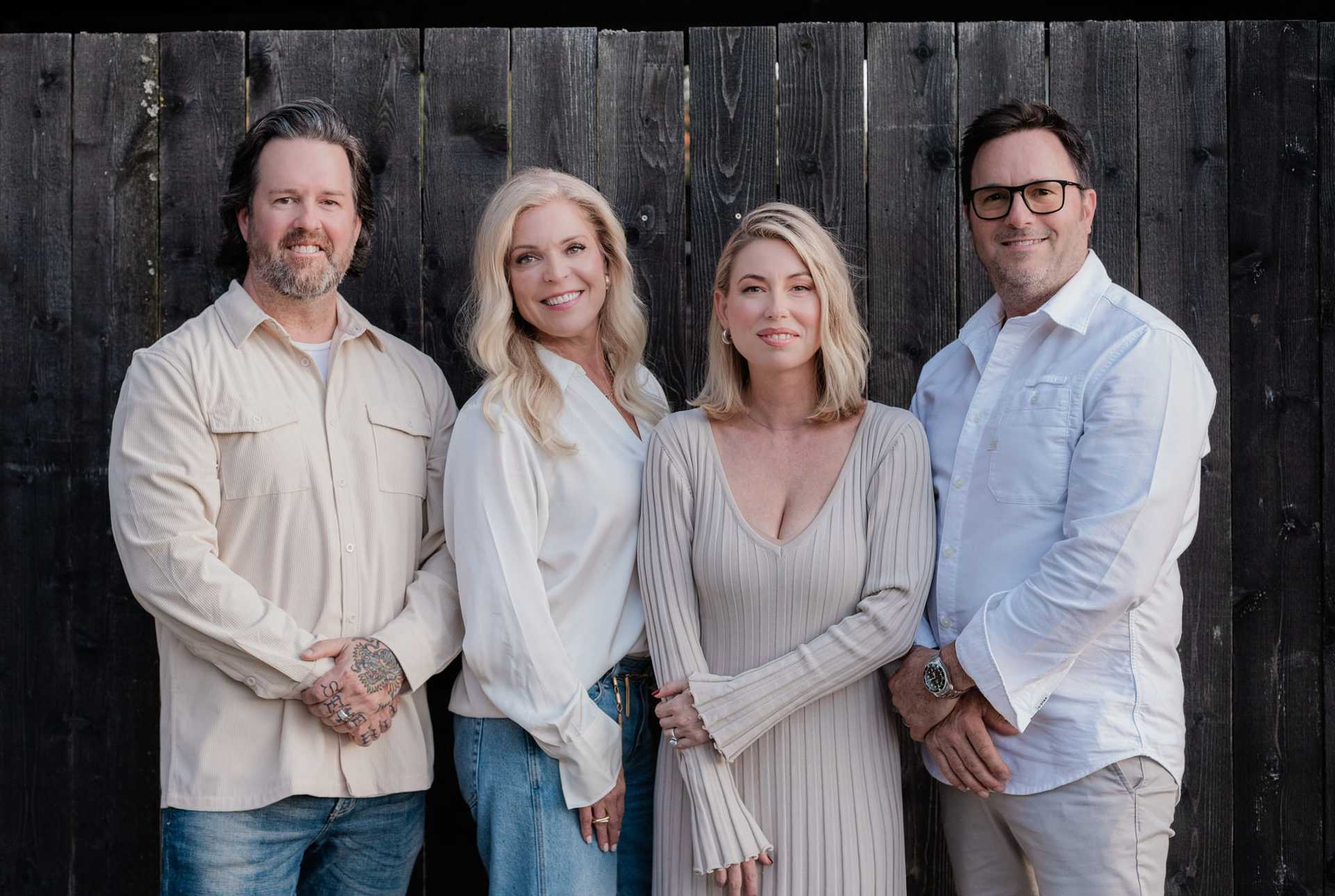 Four smiling individuals standing against a dark wooden fence, wearing light-colored clothing.