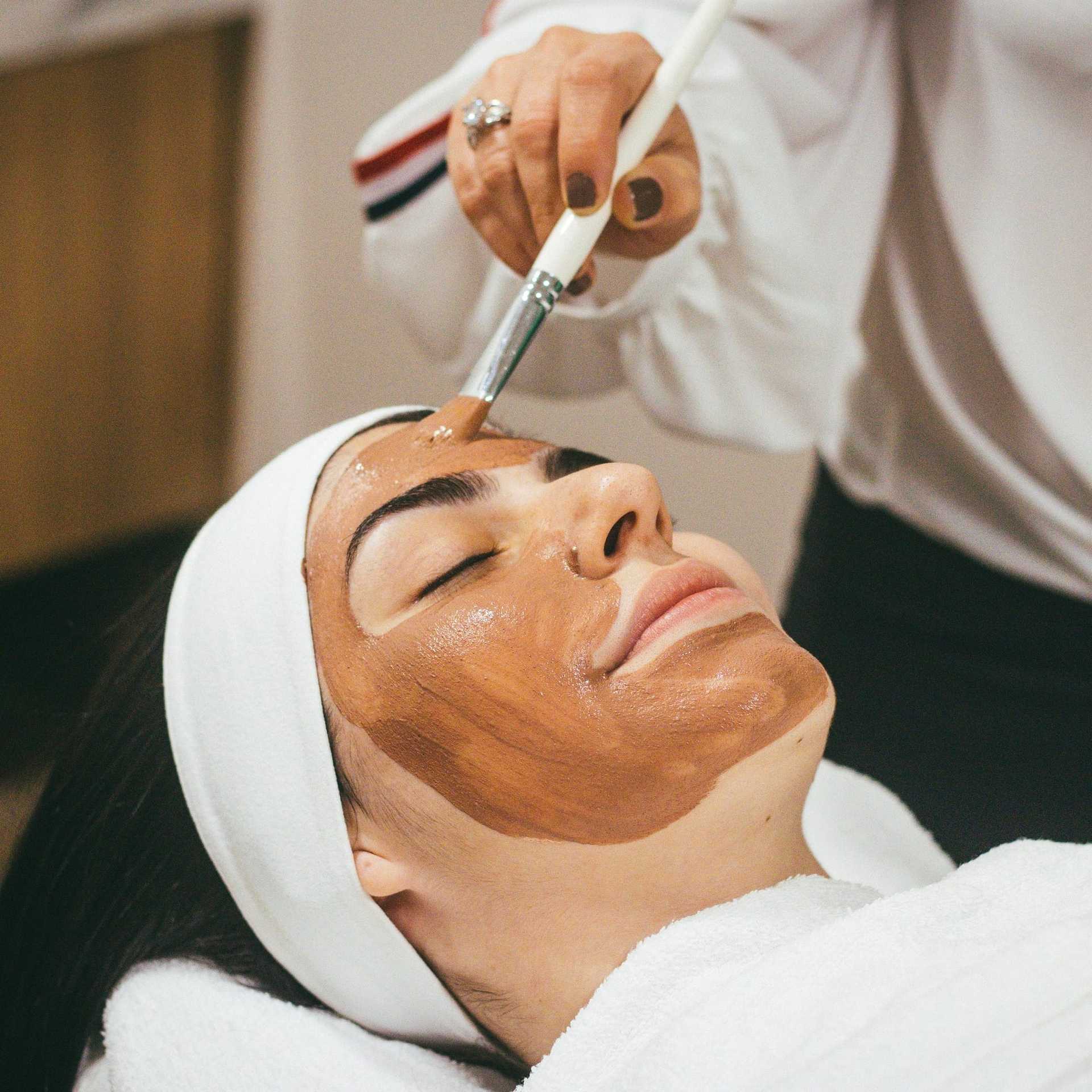 Facial treatment with a brown clay mask applied to a woman's face in a spa setting.