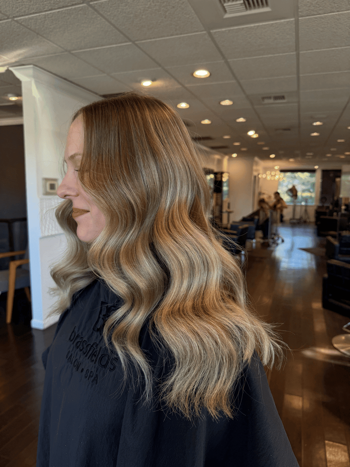 Woman with wavy blonde hair in a salon, wearing a cape.